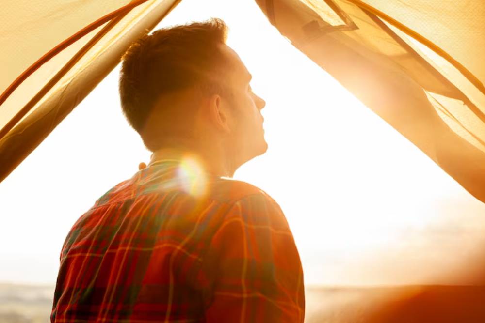 A man sits in thoughtful reflection as the sun rises outside a tent, symbolizing clarity and healing in an addiction aftercare program Columbus OH.